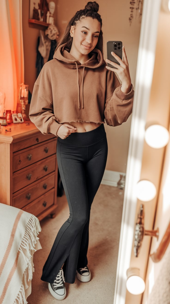 A teenage girl taking a mirror selfie in a cozy school outfit featuring an oversized gray hoodie, black flared pants, and white sneakers. The room has a modern aesthetic with a soft pink wall, a stylish bed, and neutral decor.