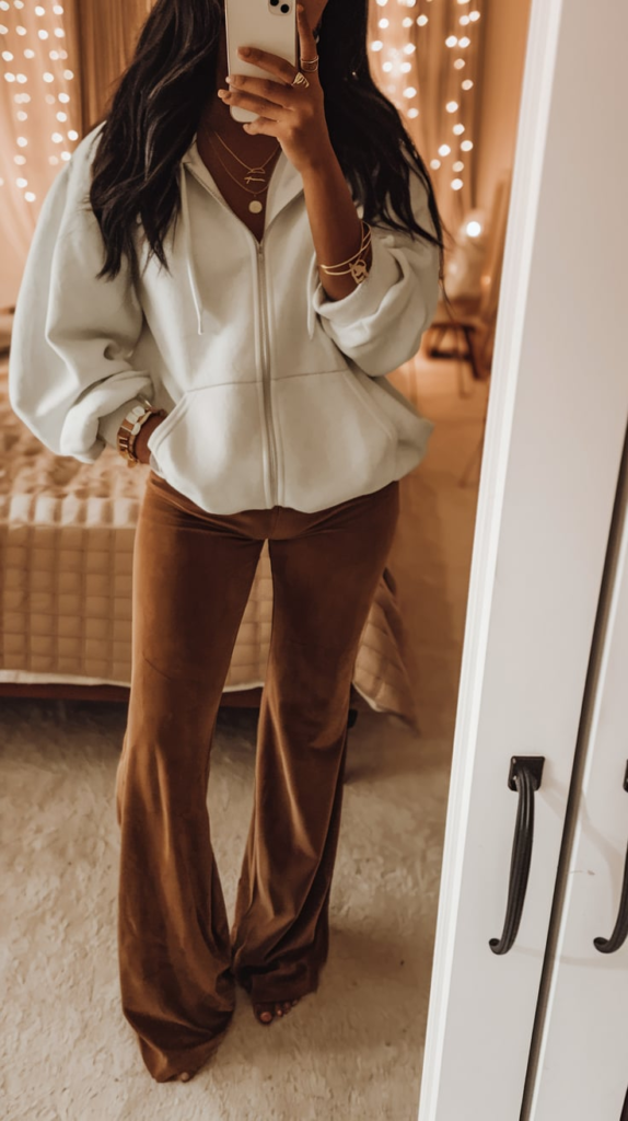 A teenage girl taking a mirror selfie in a cozy school outfit featuring an oversized gray hoodie, black flared pants, and white sneakers. The room has a modern aesthetic with a soft pink wall, a stylish bed, and neutral decor.